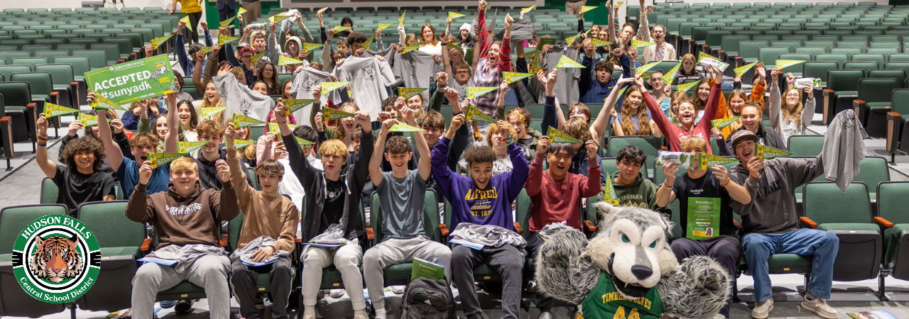 A group photo of seniors in the auditorium holding SUNY Adirondack pennants and signs for admission into SUNY Adirondack. Also pictured with Timberwolf mascot in the front