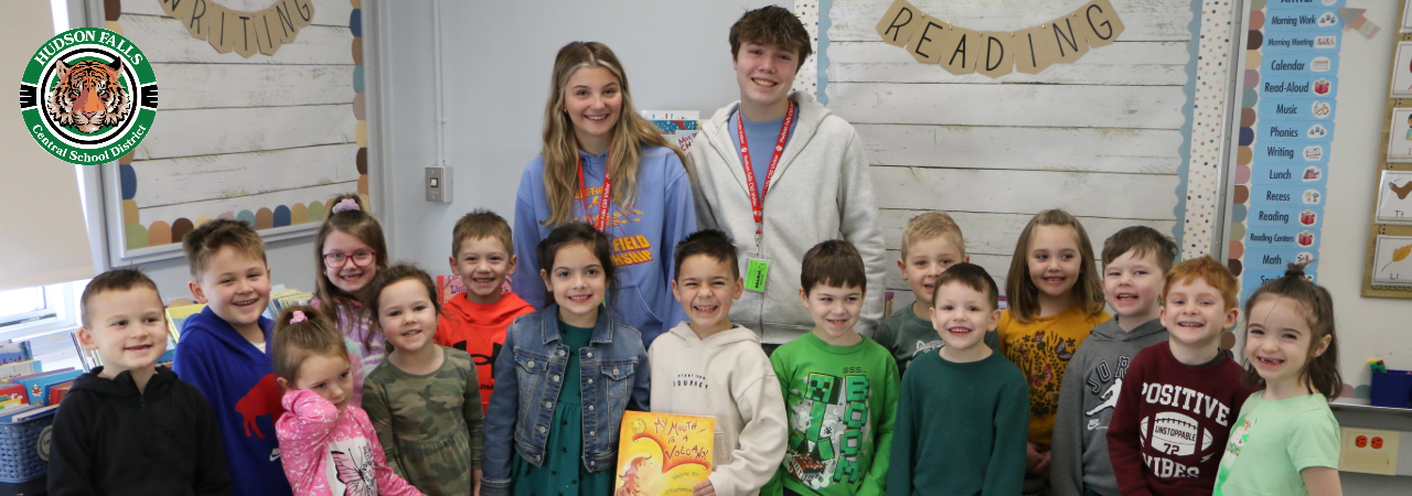 Photo of a boy and girl varsity athletes posing with a kindergarten class with a book